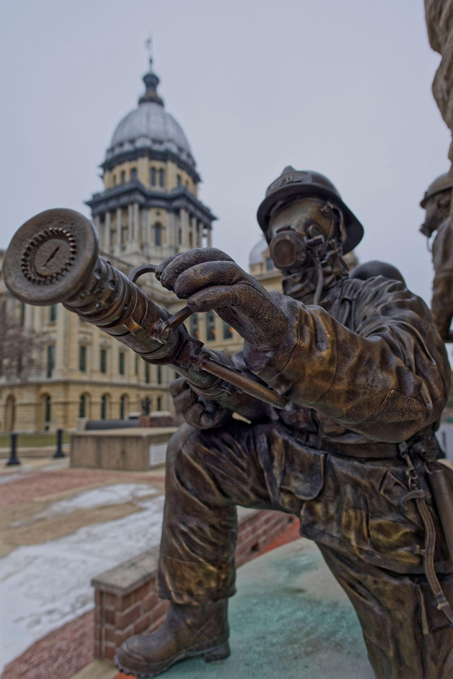 Artistic bronze firefighter statue stands in front of historic state capitol building.