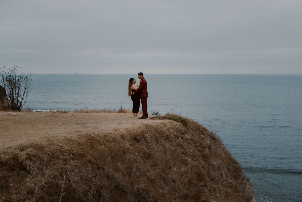 A couple in burgundy and black attire stands silhouetted on a cliff overlooking the ocean.