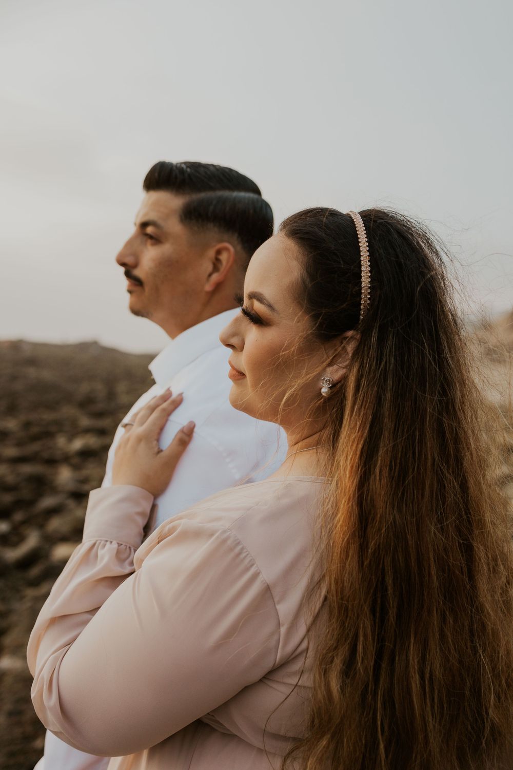 A couple in coordinated outfits stands in profile against a rocky coastal backdrop.