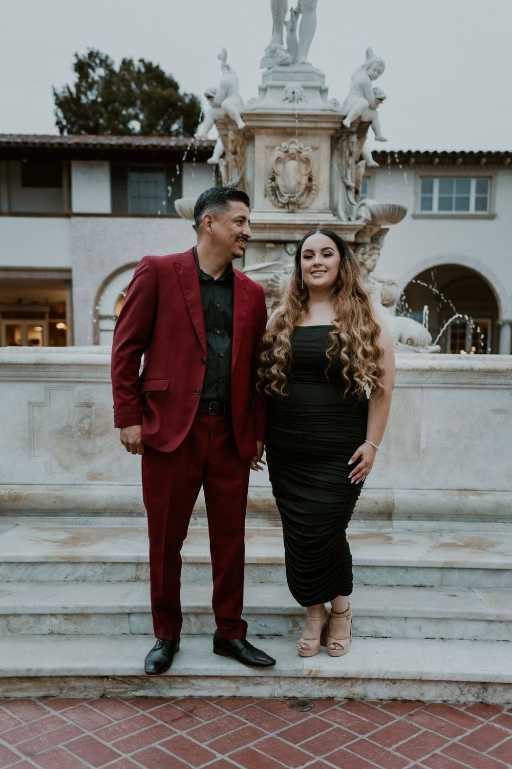 A couple in formal attire poses on stone steps in front of a decorative fountain.