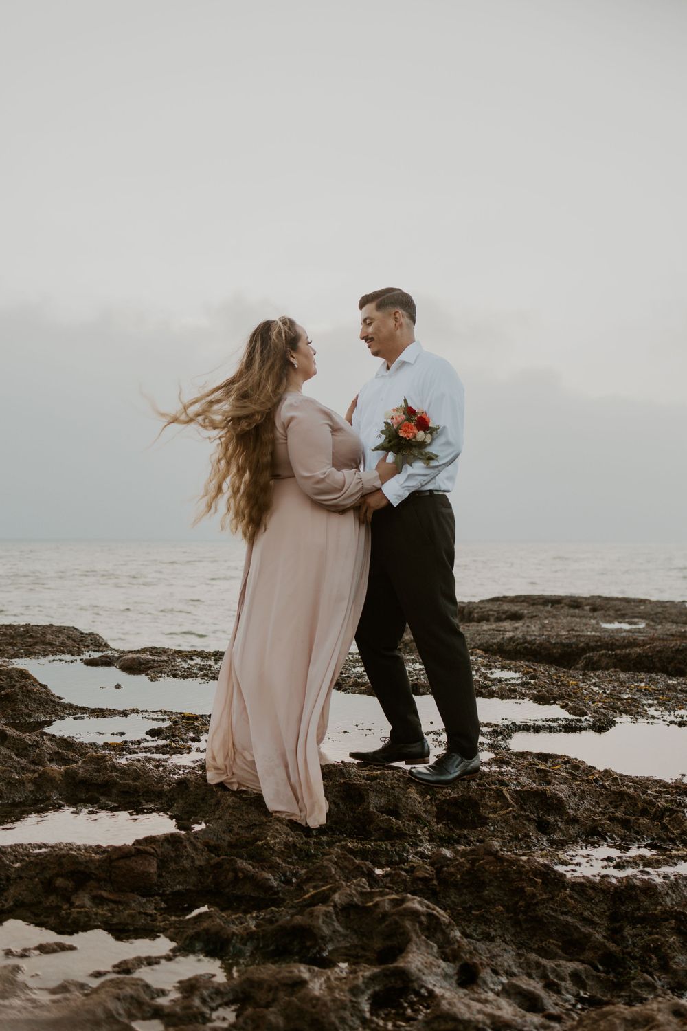 A couple shares a tender moment on rocky ocean cliffs with the woman's hair flowing in the wind.
