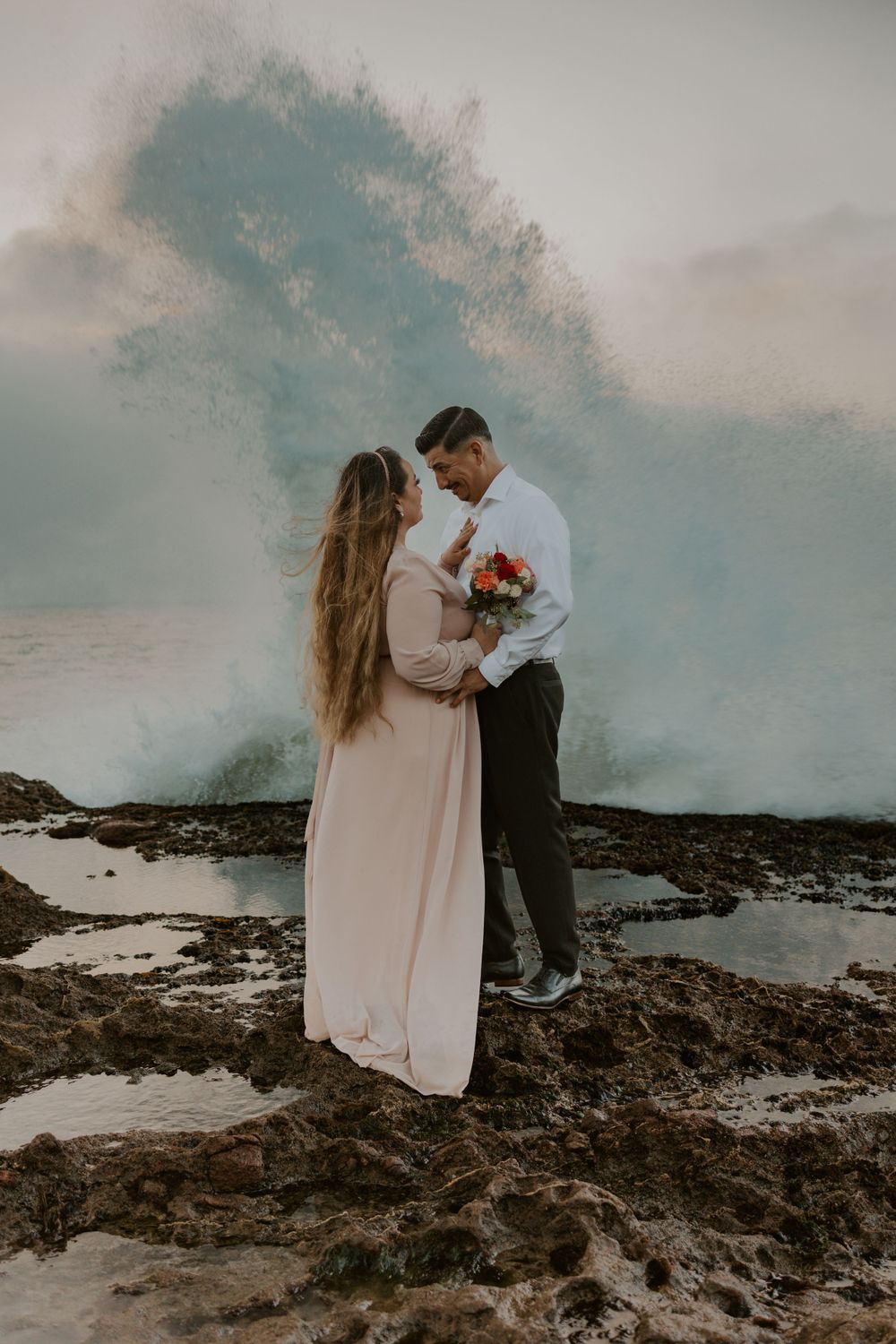 A couple embraces on rocky coastal cliffs with dramatic ocean waves crashing behind them.