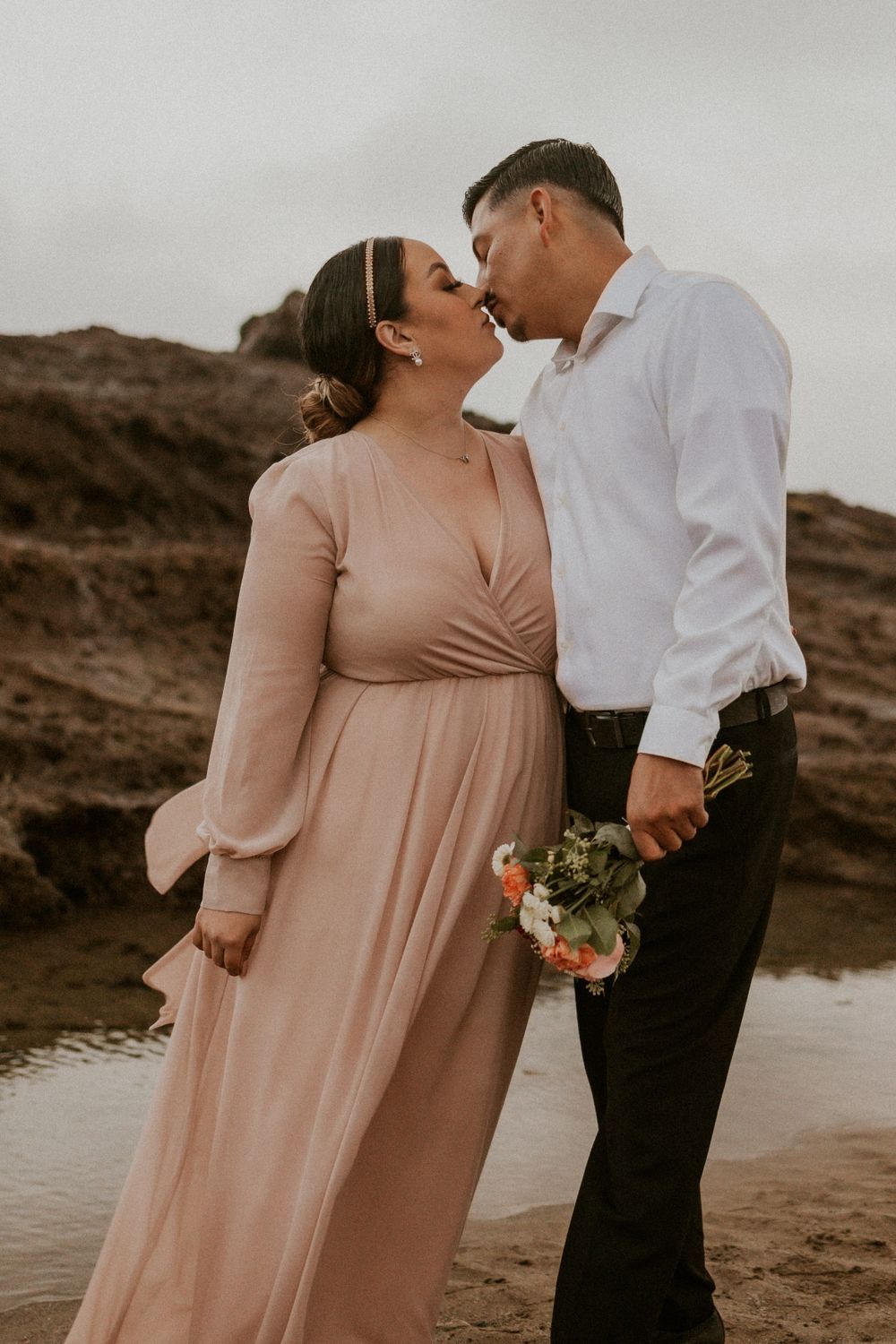 A romantic couple in formal attire share an intimate moment against a moody desert landscape.