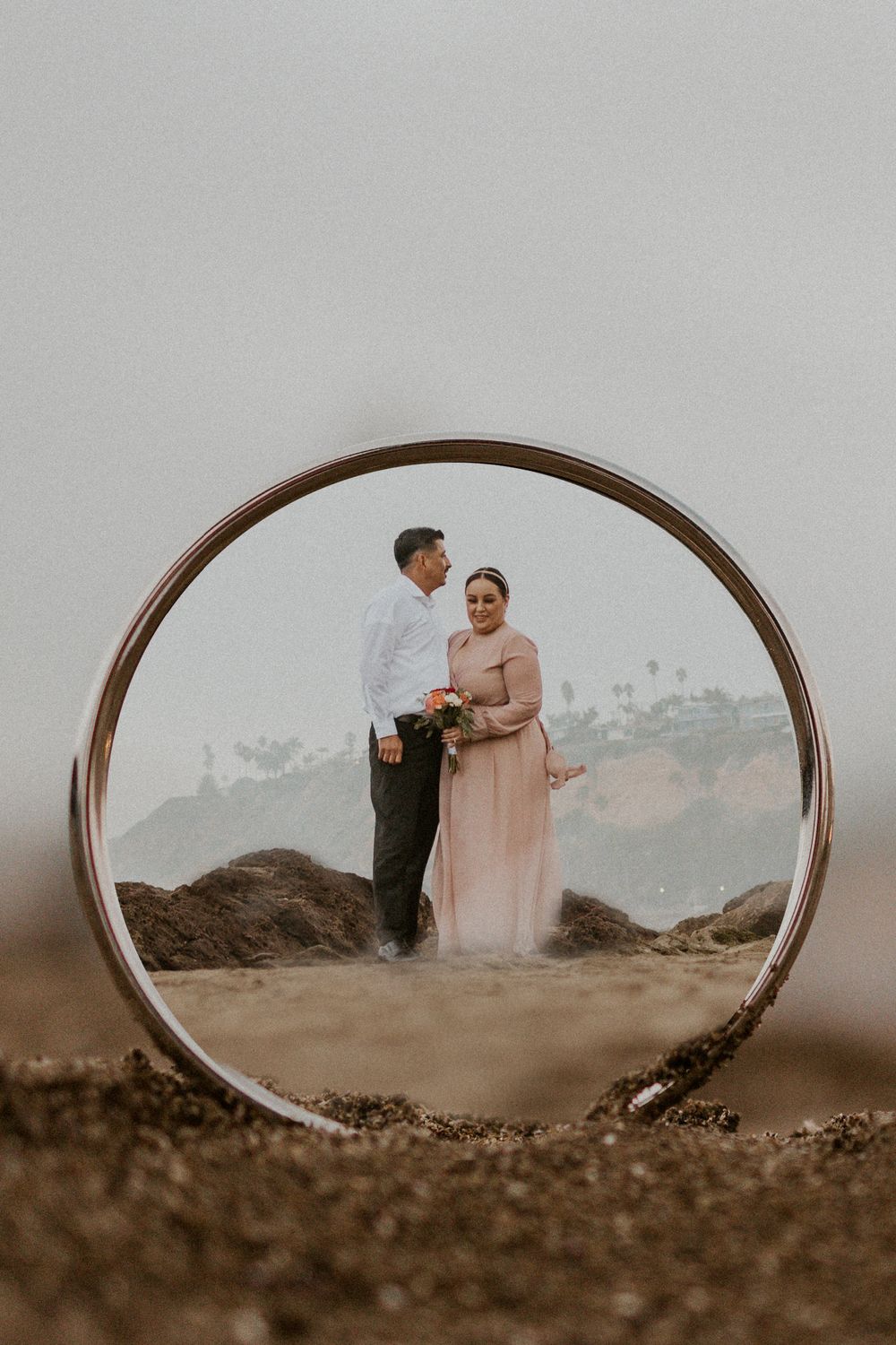 A couple poses on rocks with palm trees visible through a wedding ring frame.