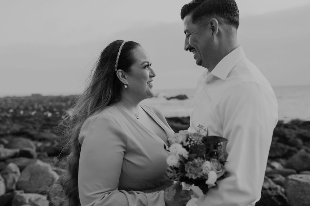 A black and white portrait of a couple sharing an intimate moment while holding a bouquet by the ocean.