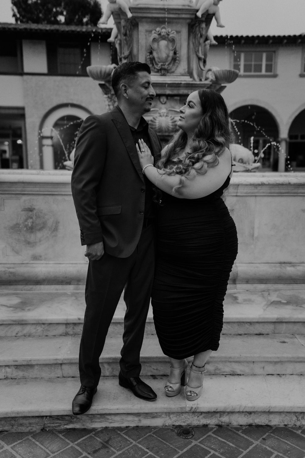 A couple poses playfully on ornate stone steps in front of a Spanish-style building.