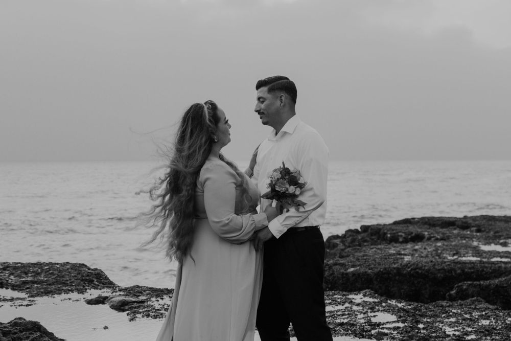 A romantic black and white beach photoshoot featuring a couple embracing on rocky coastal terrain.