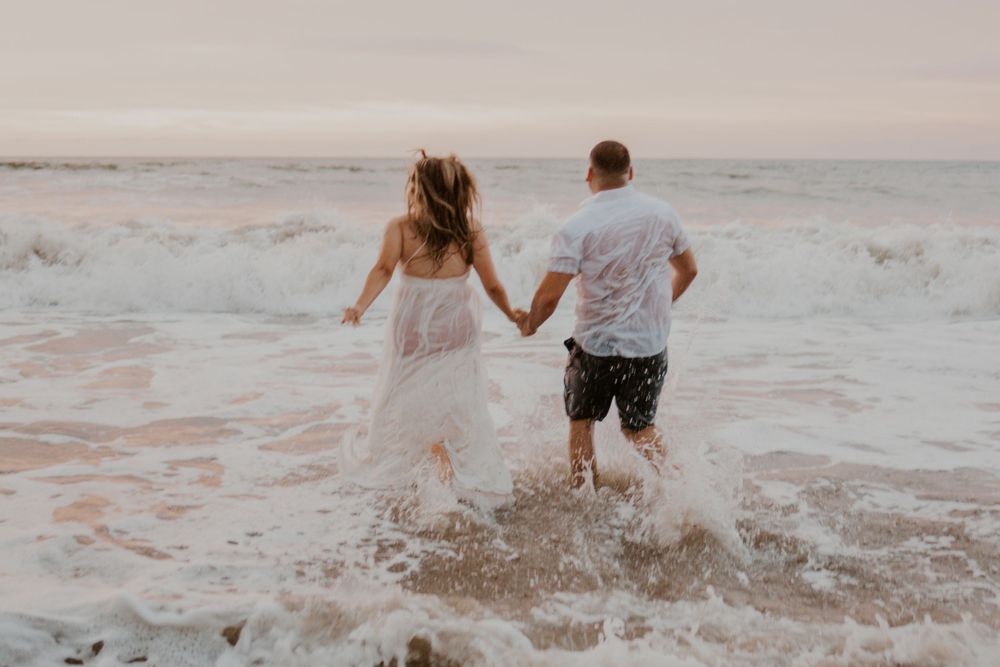 A couple splashes joyfully through ocean waves at sunset while holding hands.