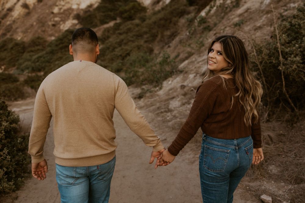 A couple holding hands while walking on a trail near scenic cliffs.