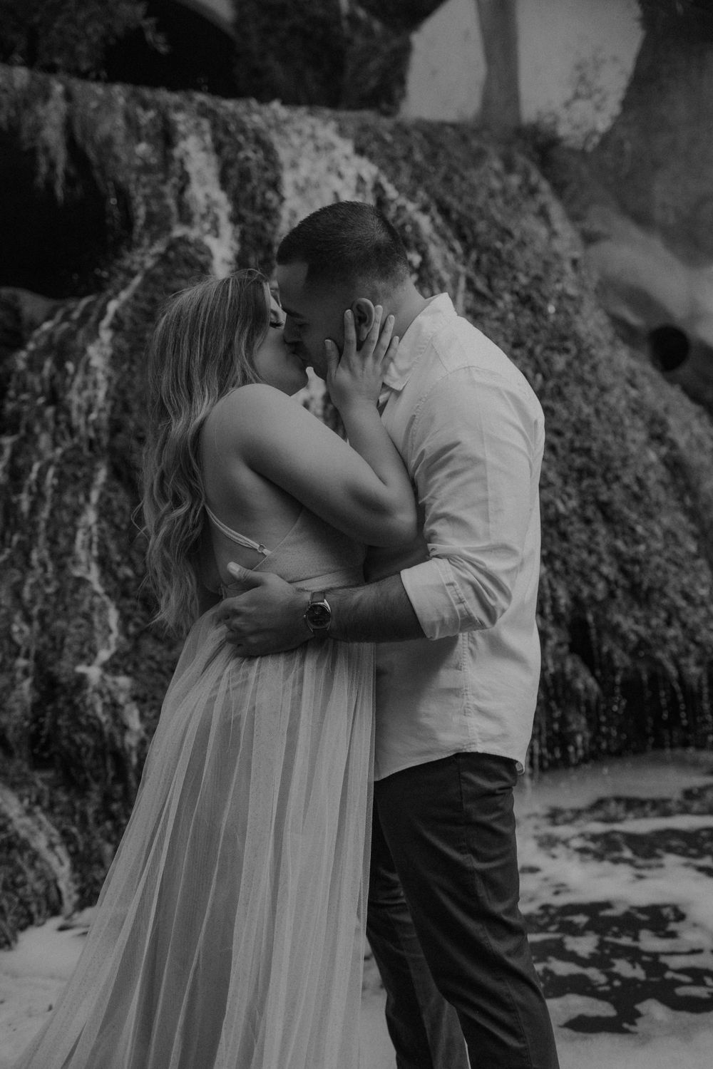 An intimate black and white portrait of a couple embracing near a waterfall.