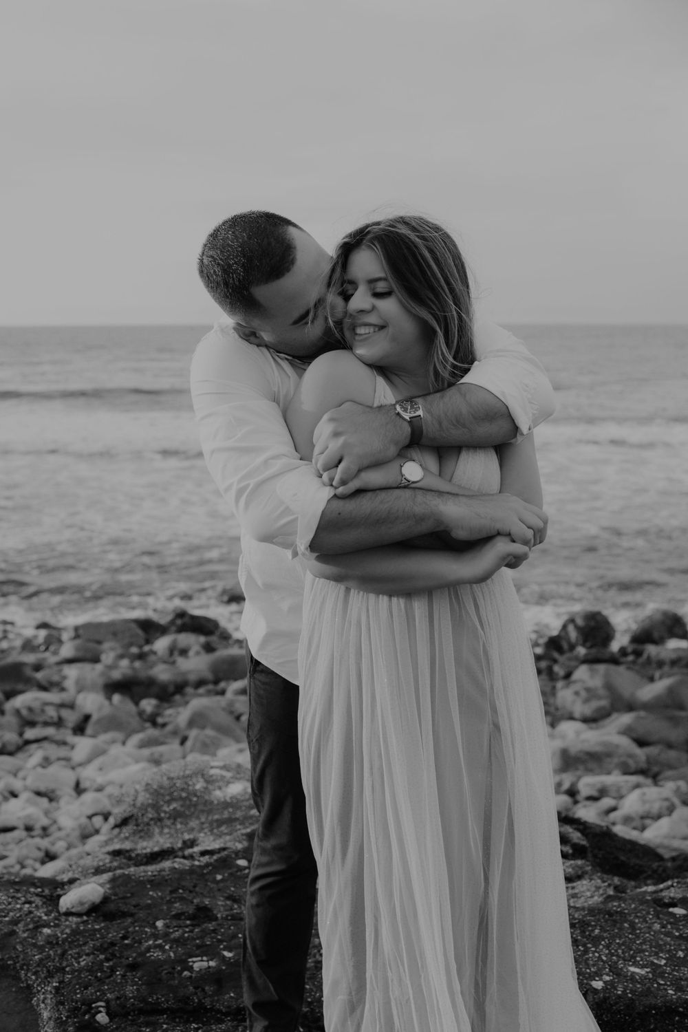 Couple in white clothing sharing tender moment by rocky oceanside.