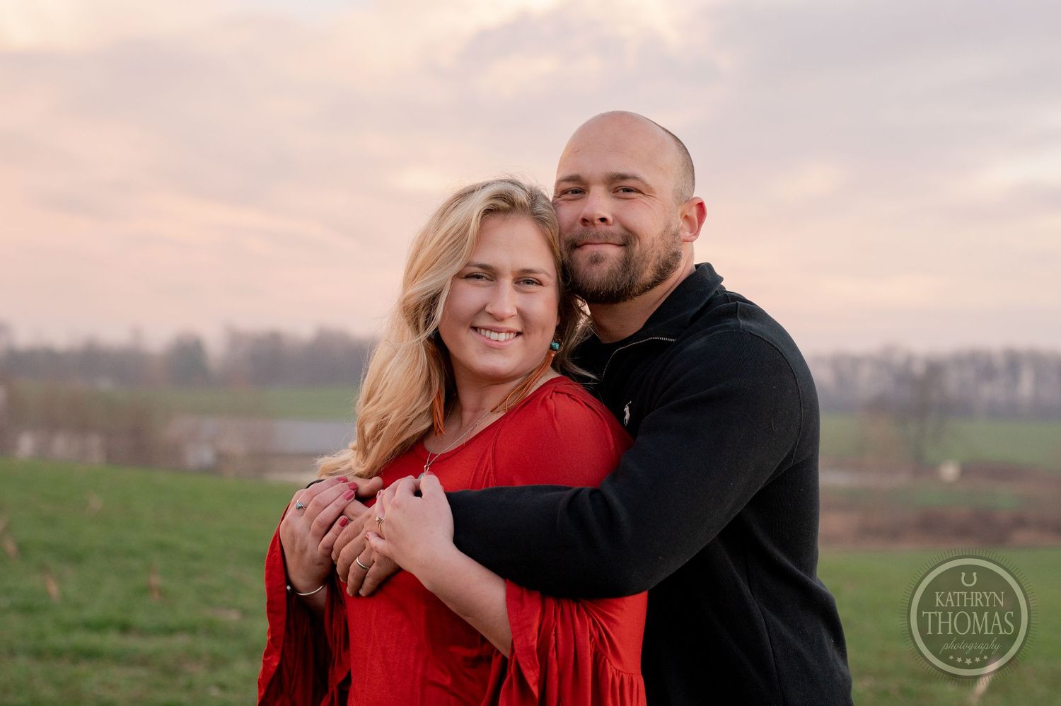 A couple embraces outdoors with warm sunset light and rolling hills in the background.