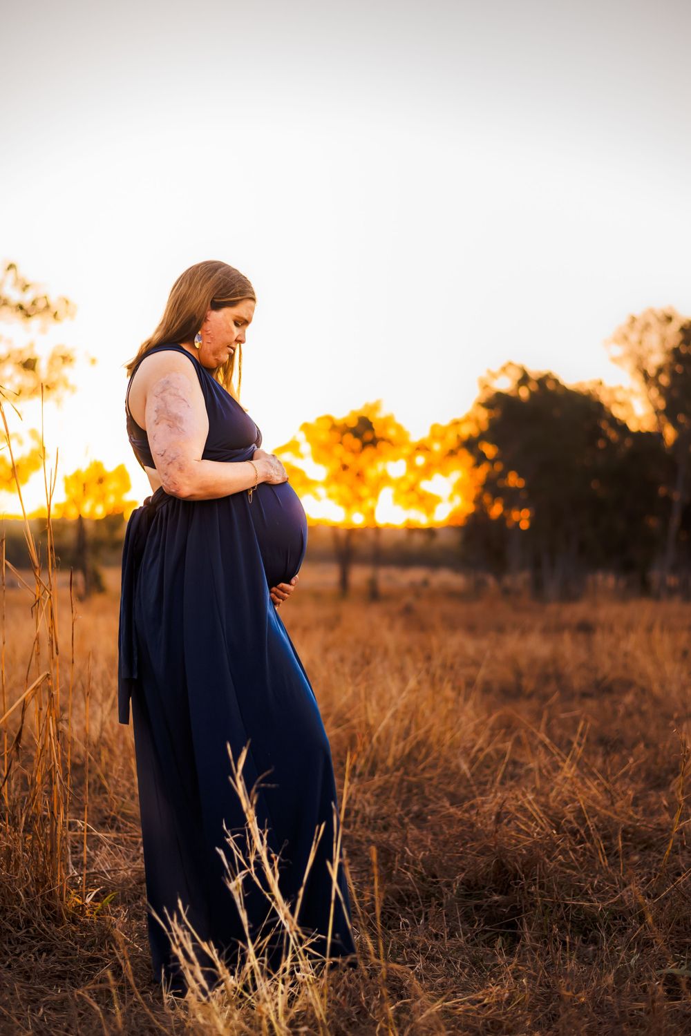 An expectant mother stands in a navy dress against a sunset backdrop in a rustic field.