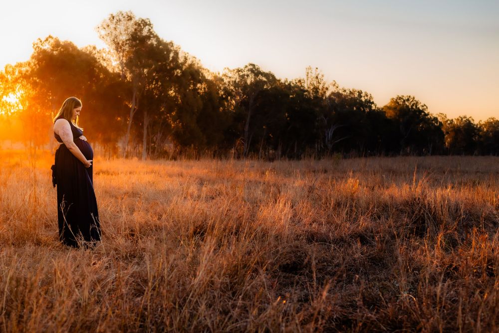 A silhouette of a pregnant woman stands in a field during golden hour sunset.