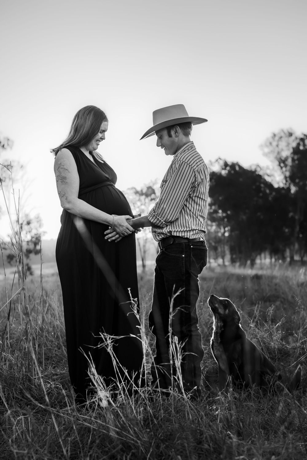 A black and white maternity photo featuring a couple and their dog in a rural setting.