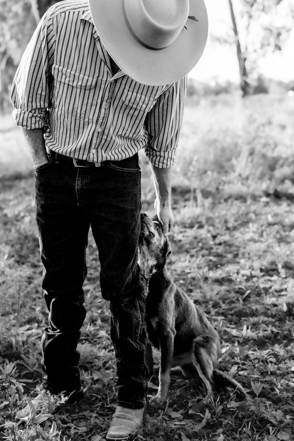 A black and white photo of someone in a cowboy hat standing with their loyal dog.