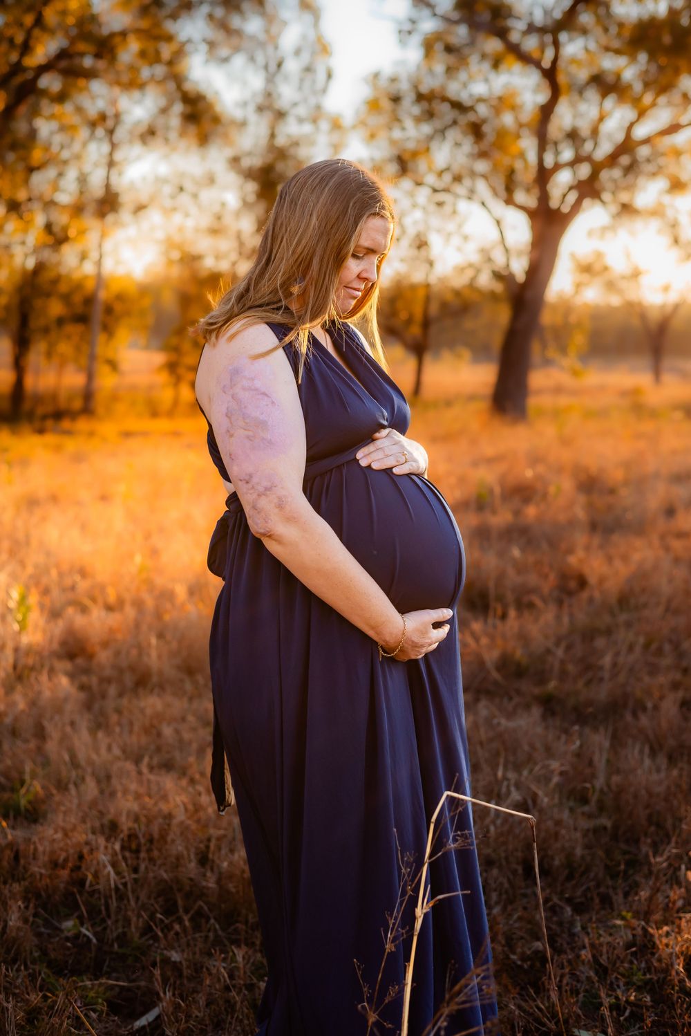 Pregnant woman in navy dress cradles belly during golden hour sunset maternity photoshoot in rural field.