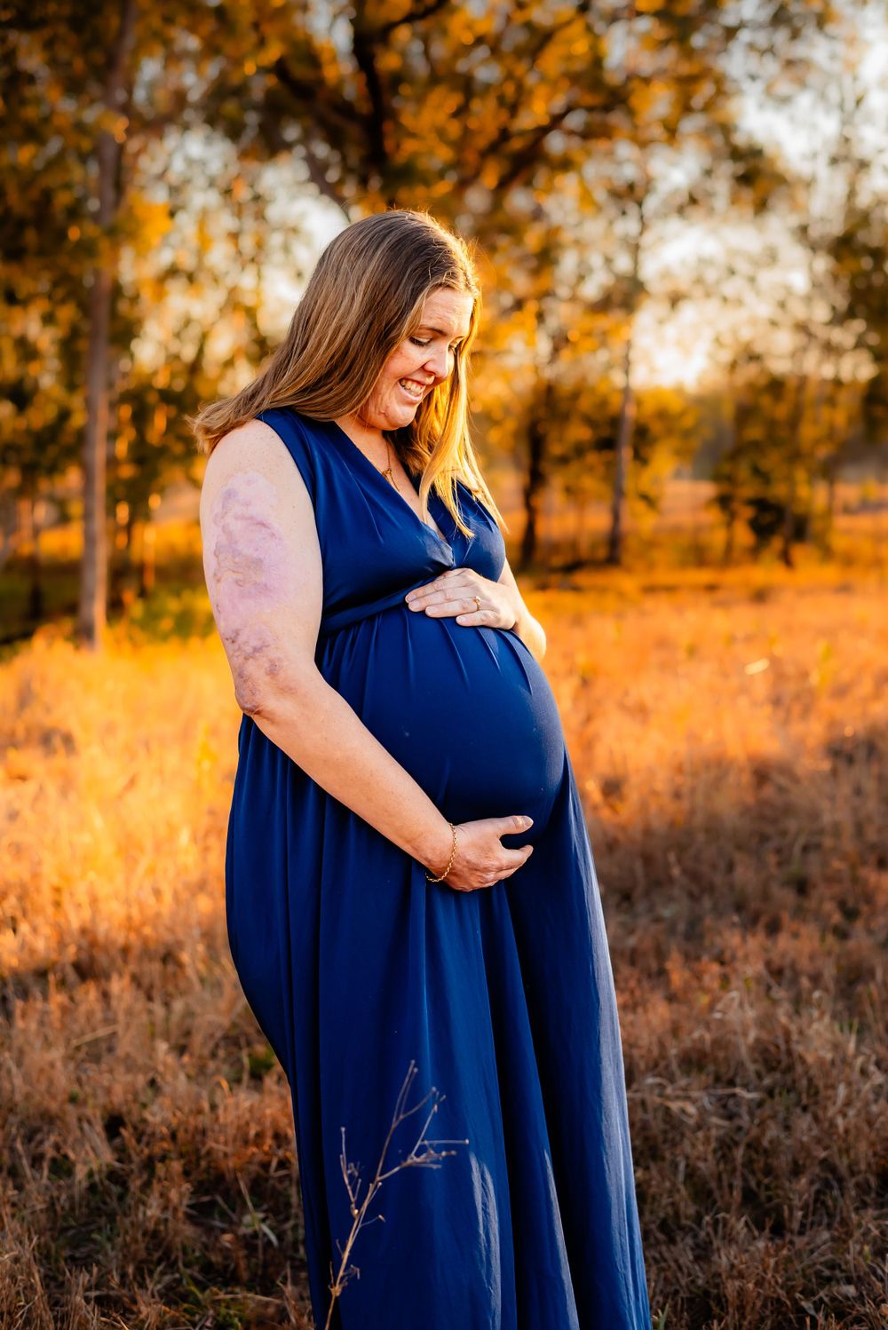 Expectant mother wearing blue maxi dress poses in autumn field at sunset.