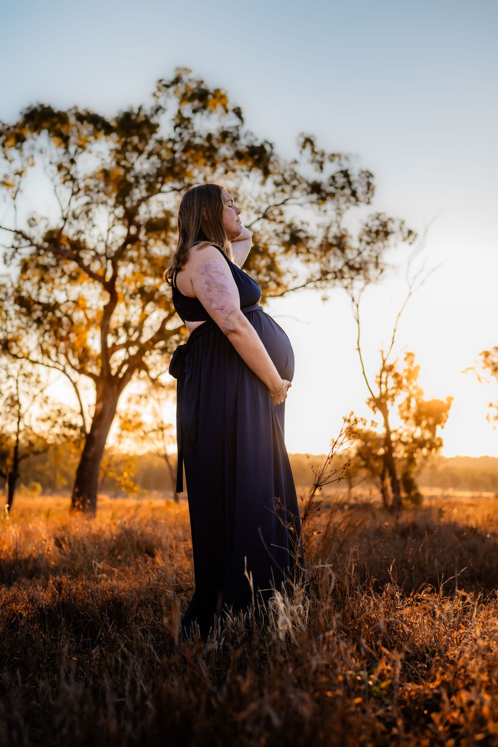 Side view of pregnant woman in dark dress against sunset in countryside setting.