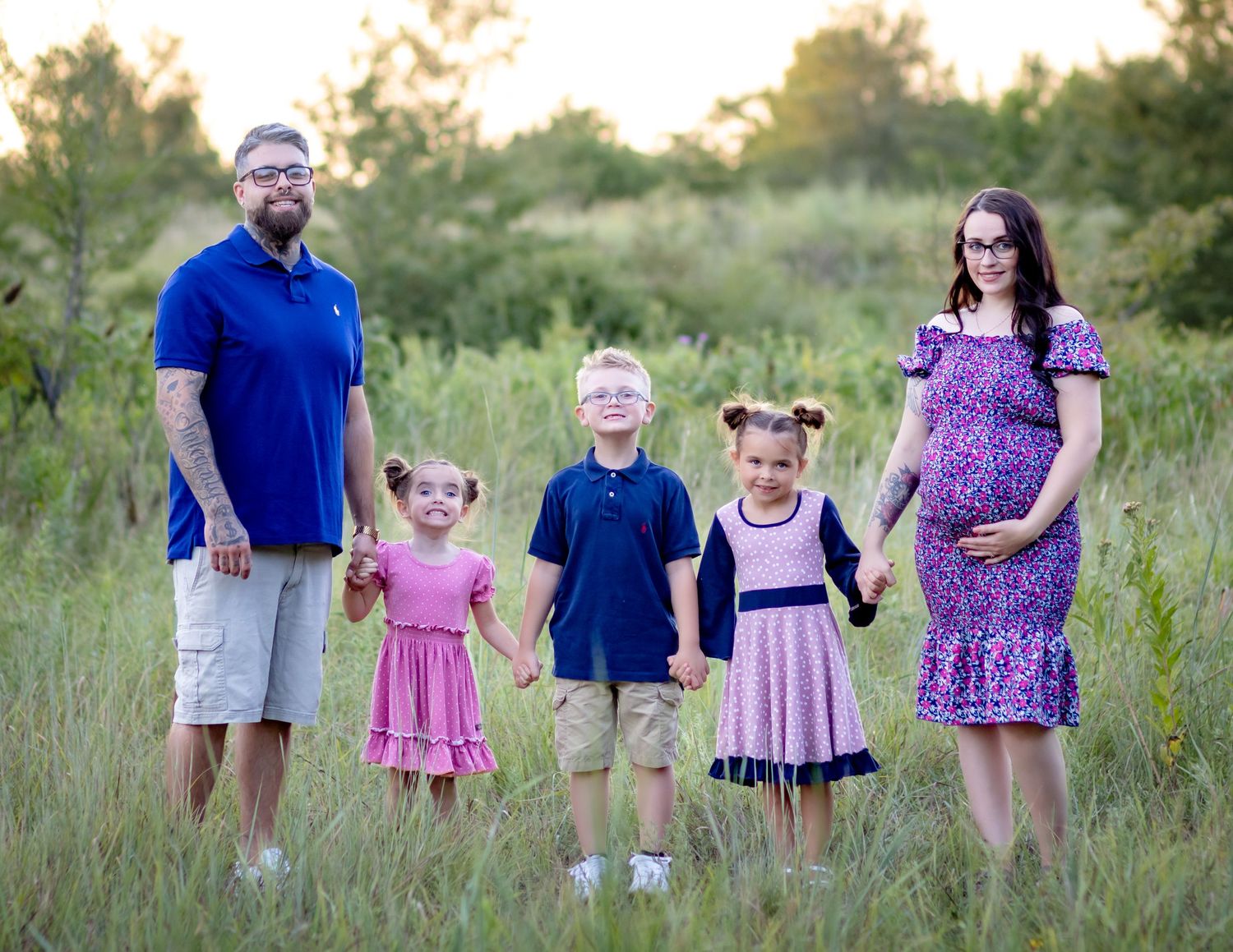 A family wearing color-coordinated outfits poses together in a sunlit field of tall grass during golden hour.