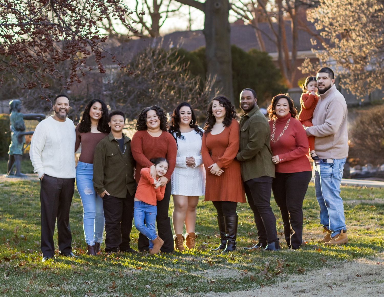 A group of people in fall attire standing together outdoors on grass during sunset with autumn trees in background.