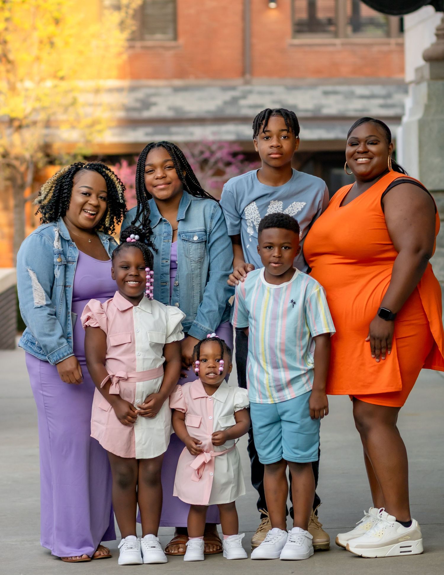 A group wearing coordinated outfits in denim jackets, purple, pink and orange gather together outside a brick building.