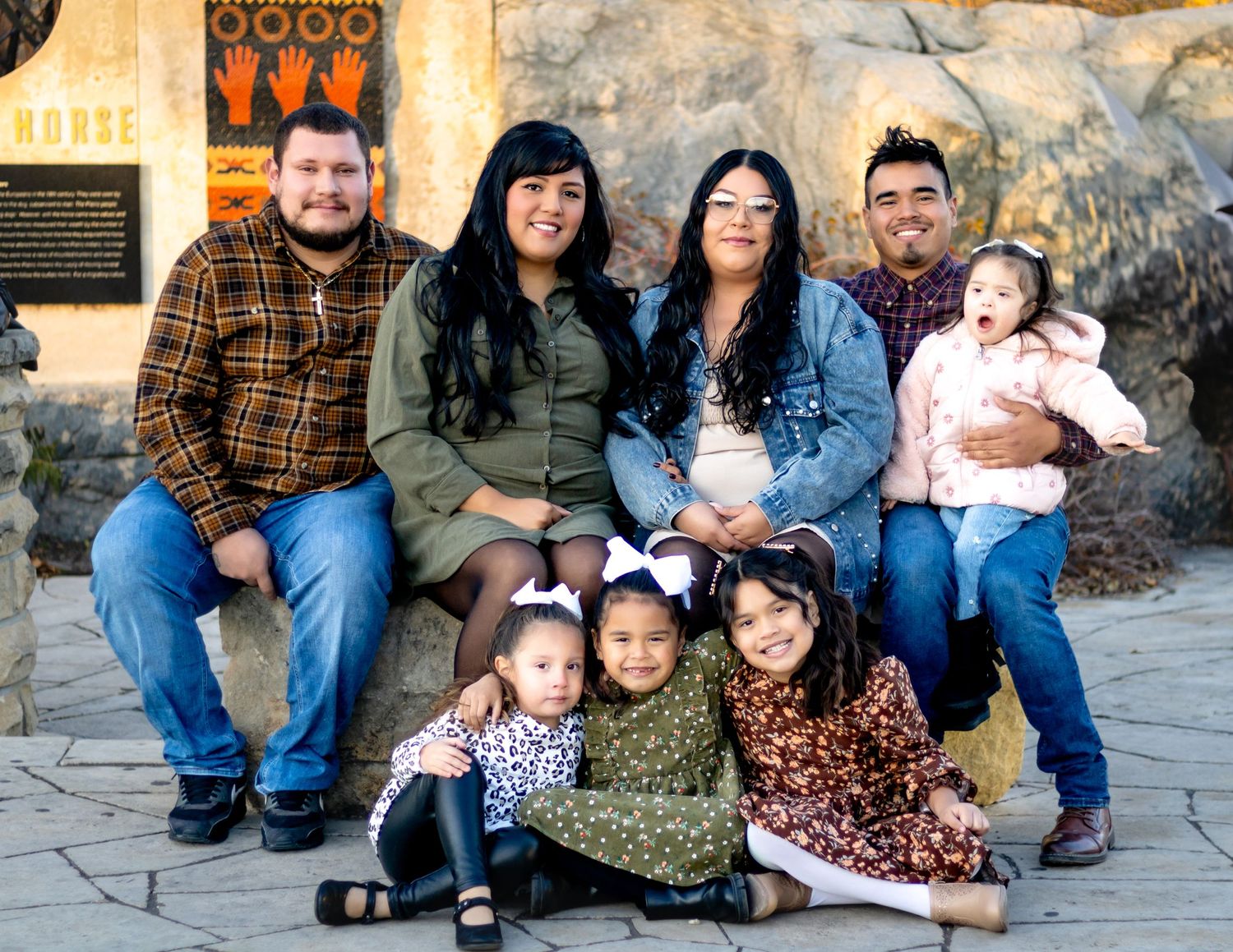 A group of adults and children pose together in casual attire against a rustic stone wall.