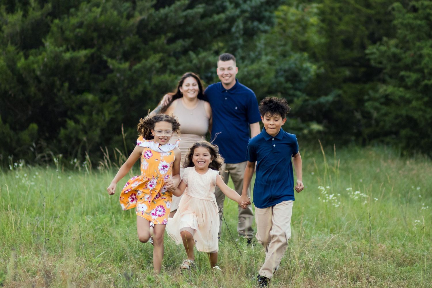 Family group running and smiling together in a green summer meadow with evergreen trees in the background.
