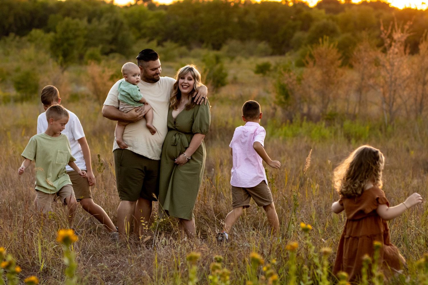 A family walks together through a golden field of tall grass and wildflowers at sunset.