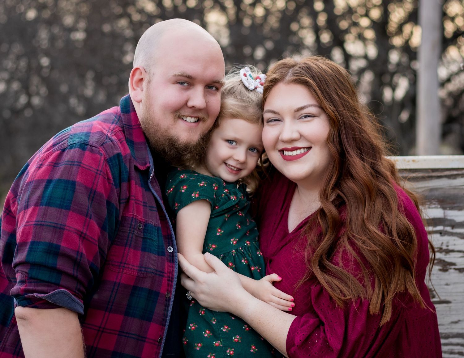 A family of three poses outdoors wearing red plaid and dark green, smiling warmly at the camera.