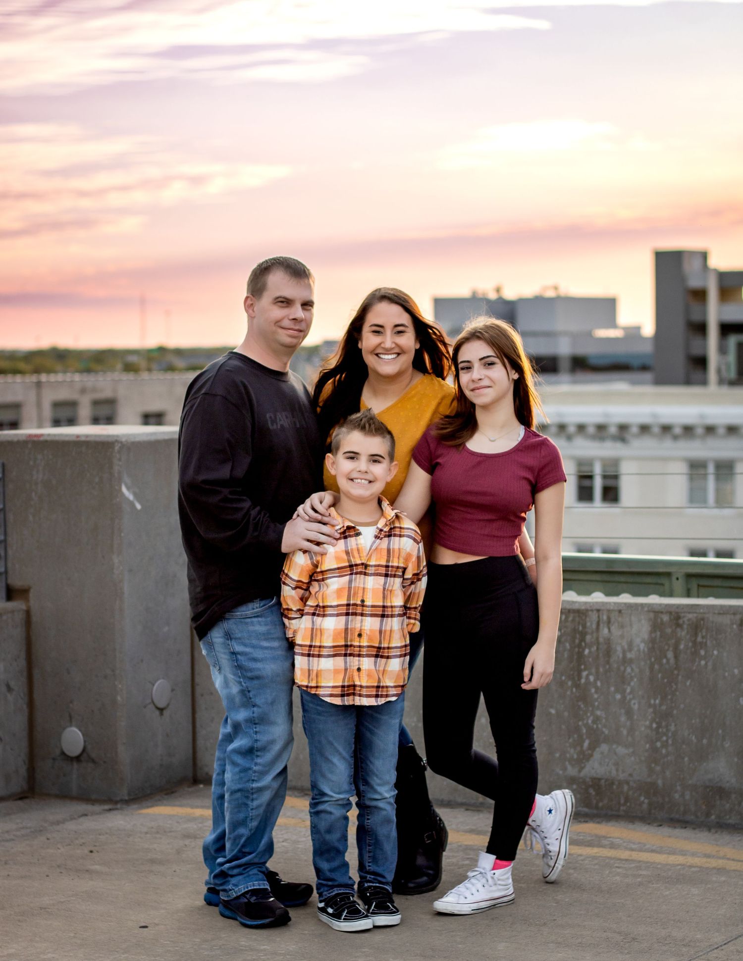 A family poses together on a rooftop during sunset with pink sky and urban buildings in the background.