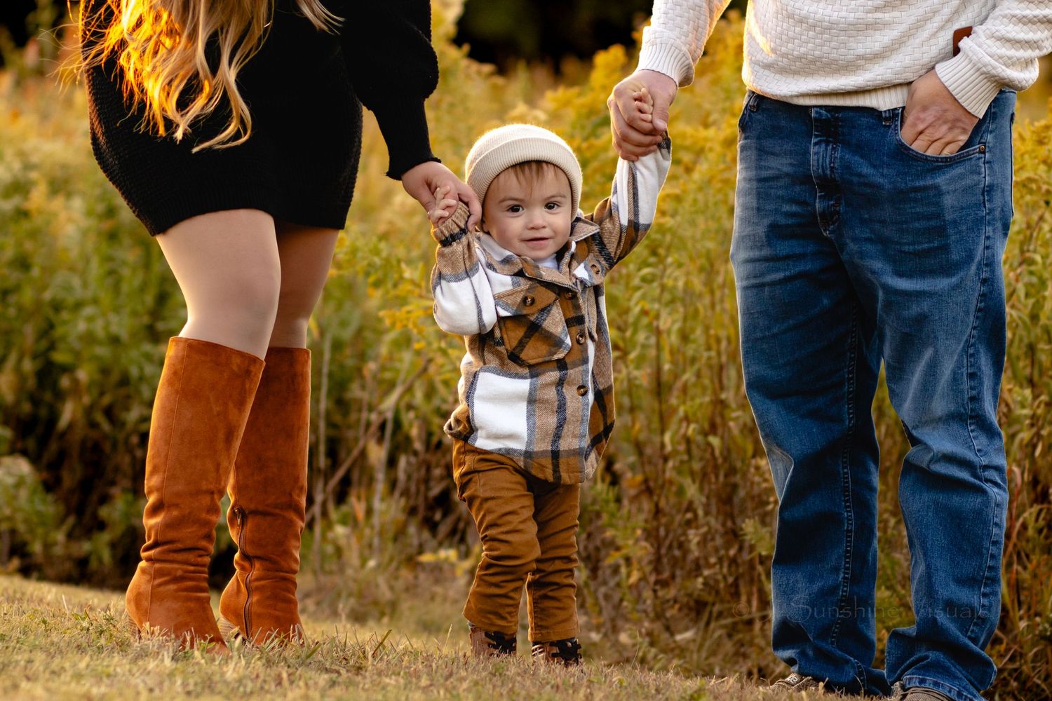A small figure in brown corduroy pants walks between two people in tall golden grass during autumn.