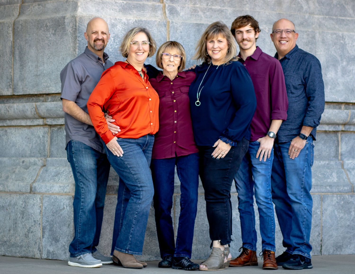 A group of six people wearing casual clothing stands together against a gray stone wall outdoors.
