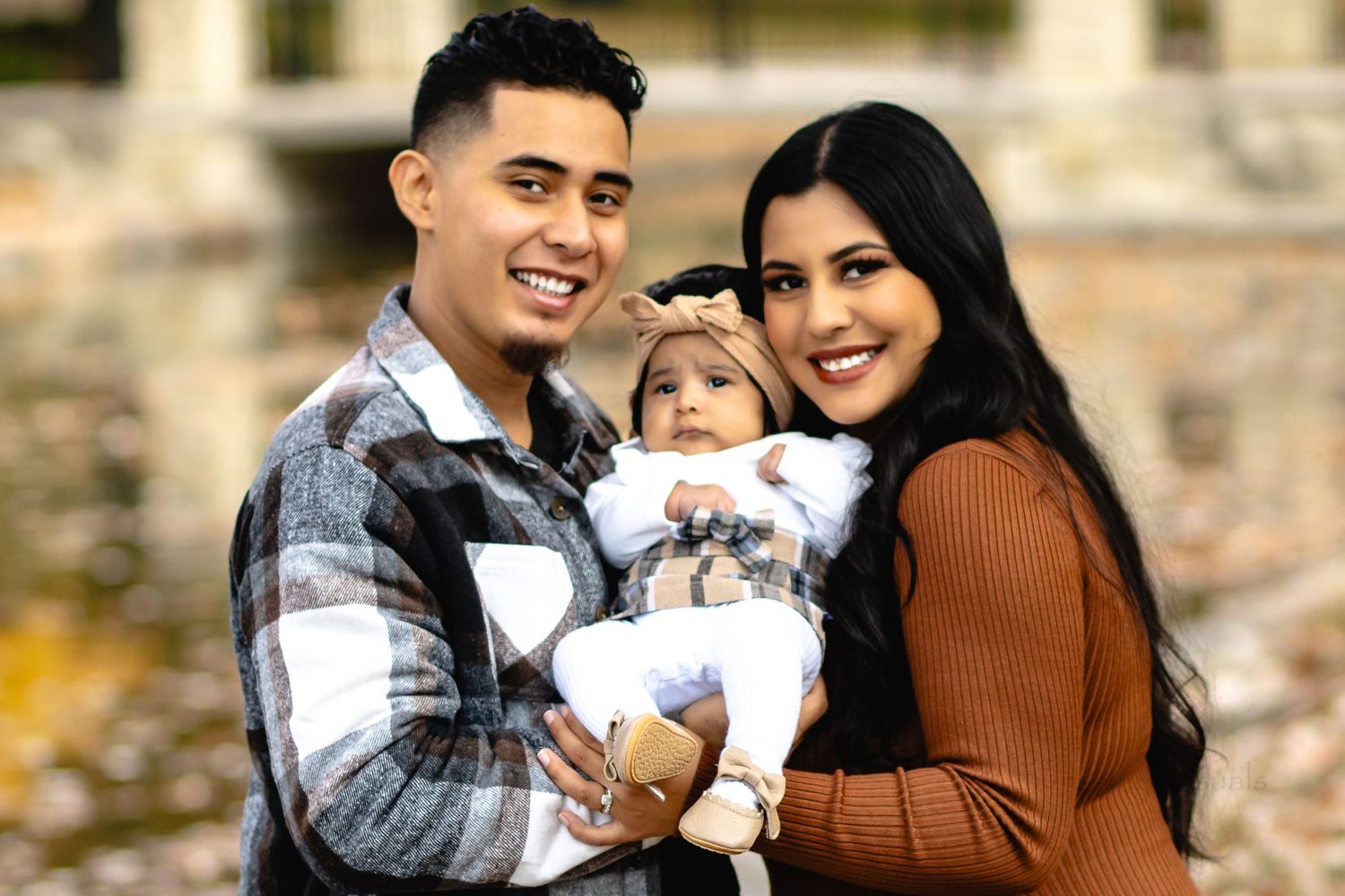 Young couple holding their baby in white clothing while posing outdoors during an autumn family photo session.