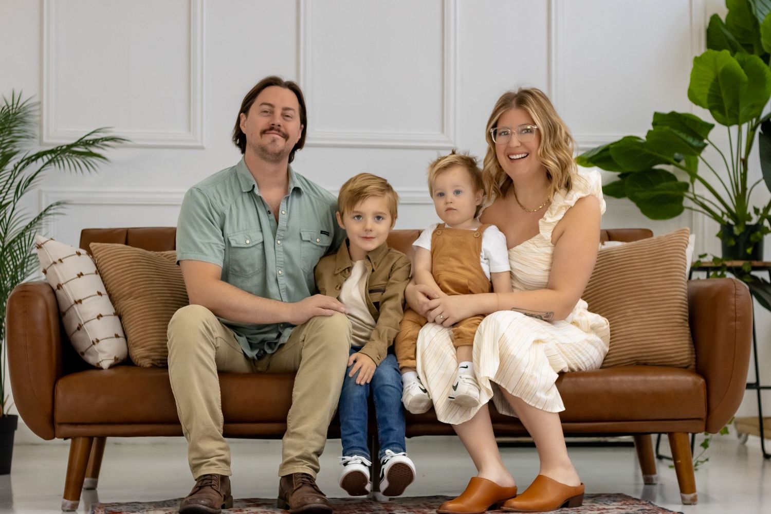 A family of four sits together on a brown leather couch with indoor plants in the background.