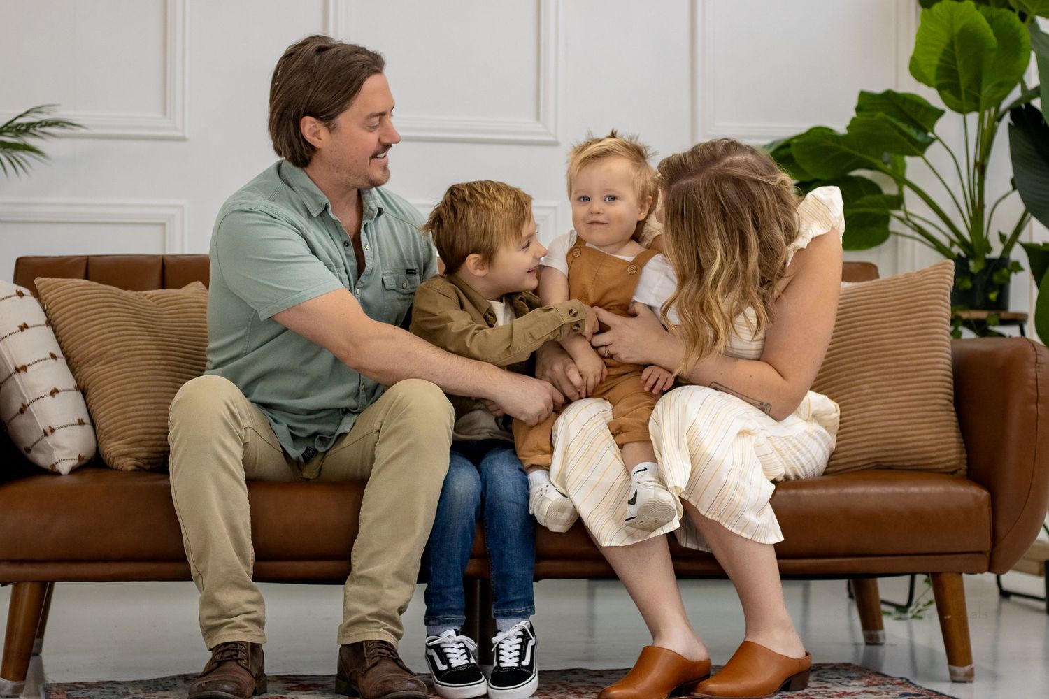 A family sits together on a brown couch in their living room with houseplants visible in the background.