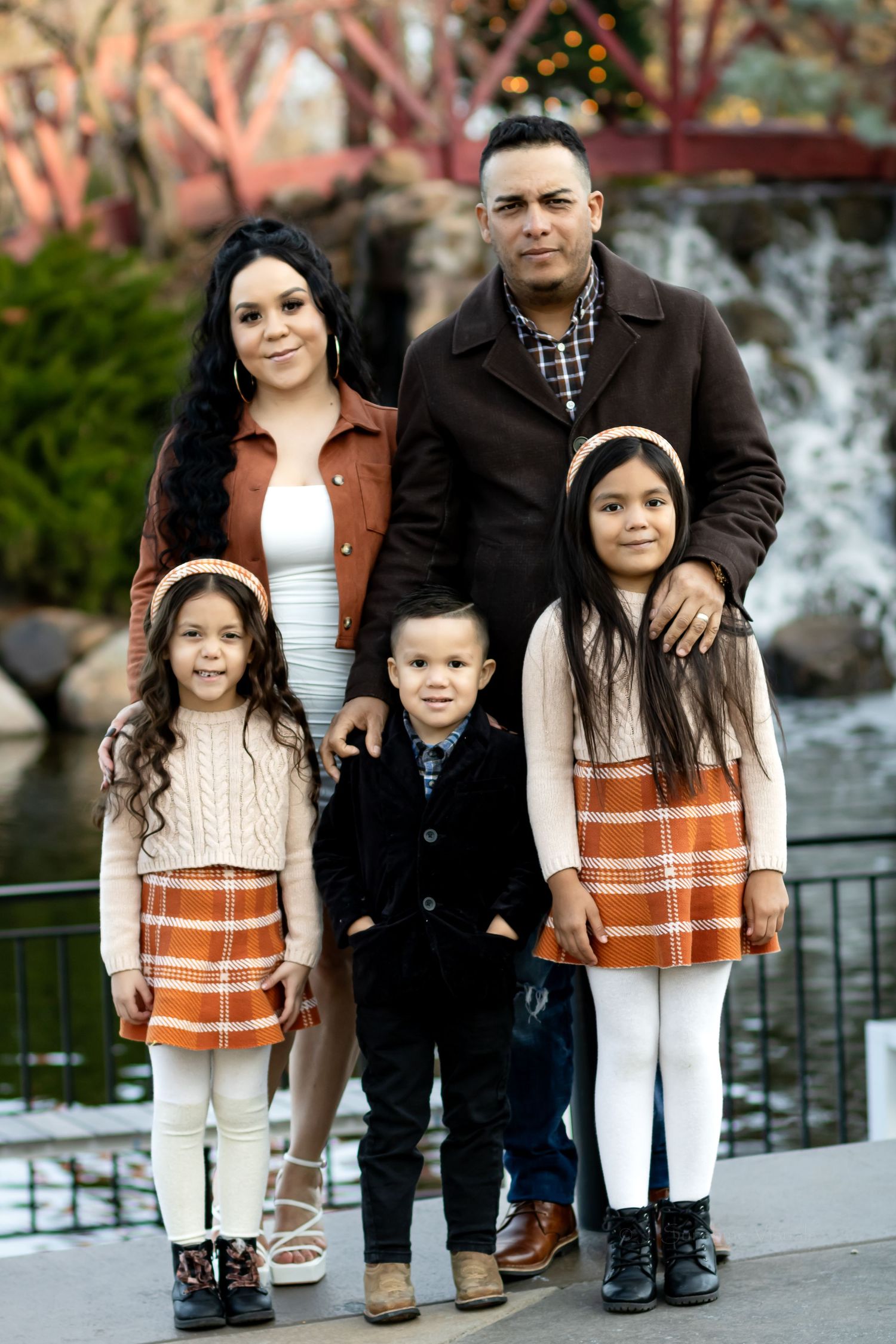 Family of five dressed in coordinating autumn outfits standing together outdoors near railing with waterfall in background.