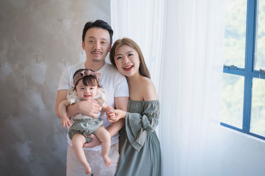 Happy young Asian family with baby posing for portrait near bright window in elegant studio setting幸福的年輕亞洲家庭帶著寶寶在優雅的攝影棚裡，在明亮的窗戶旁擺姿勢拍攝肖像照。