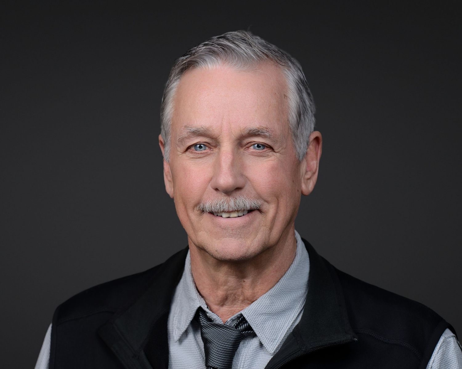 Professional headshot of senior man in suit and tie against dark background.