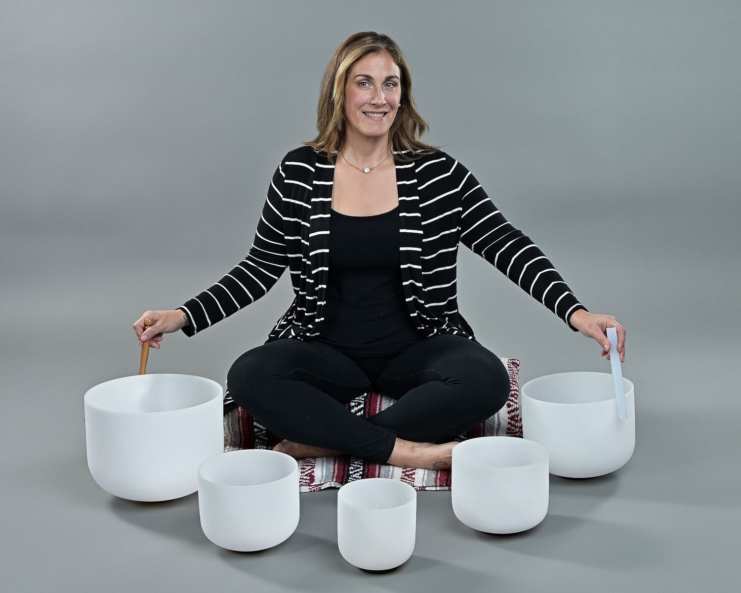 A person sits cross-legged on the floor surrounded by white crystal singing bowls against a gray studio backdrop.