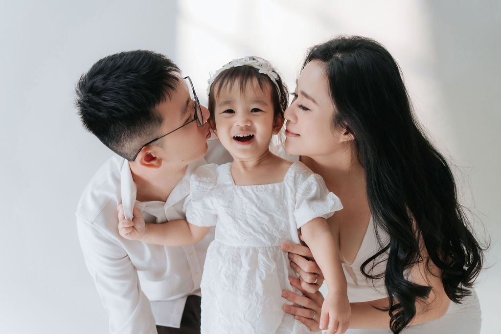 Happy family portrait with parents kissing their laughing toddler daughter in white outfits against a bright background.幸福的全家福，父母親吻著身穿白色衣服