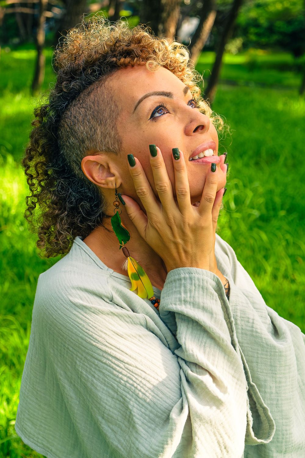 Woman with curly hair and green nails covers mouth in surprise, smiling outdoors in lush green park setting.