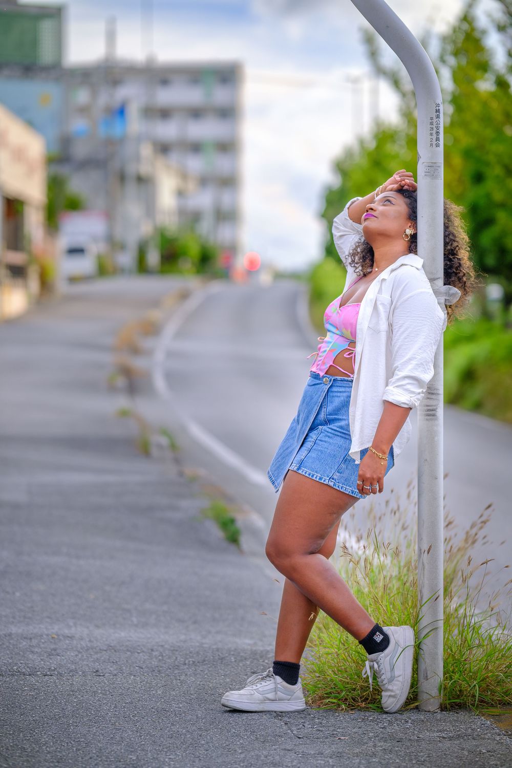 A person in denim shorts and white top poses casually against a street pole in an urban setting.