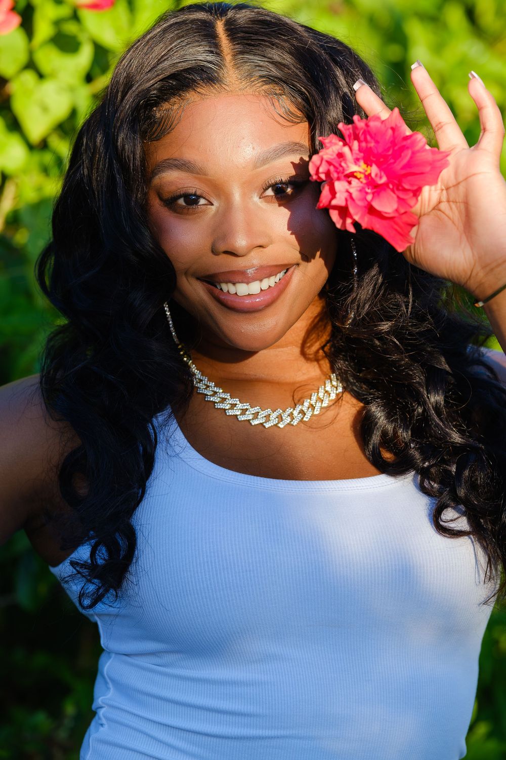 Smiling person in light blue tank top holds pink flower while wearing silver chain necklace against green foliage background.