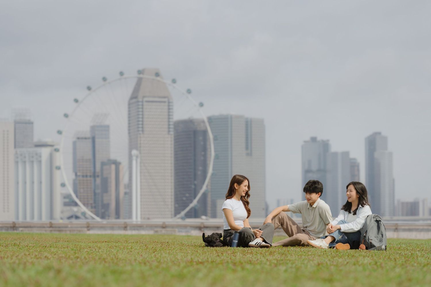 Three friends sit on green grass with Singapore Flyer and city skyline in the background.