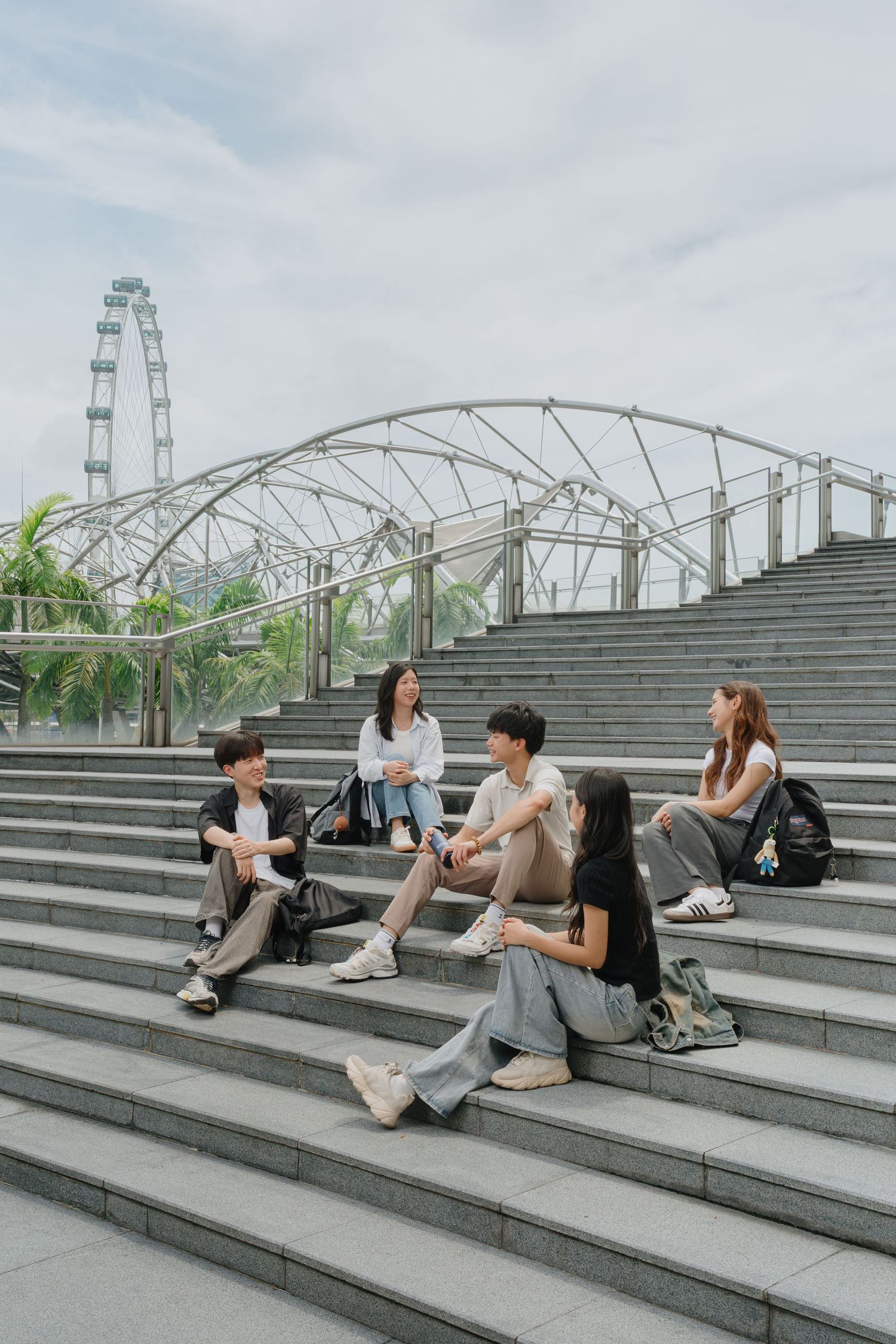 Five young adults sit on outdoor steps near the Helix Bridge and Singapore Flyer, chatting casually.