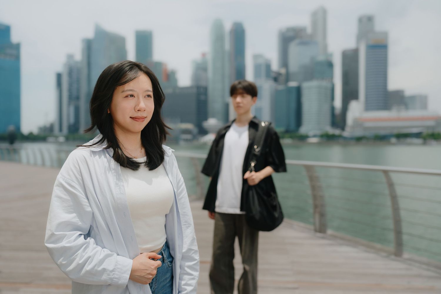 Woman in white jacket stands on waterfront promenade with Singapore city skyline and man in background.