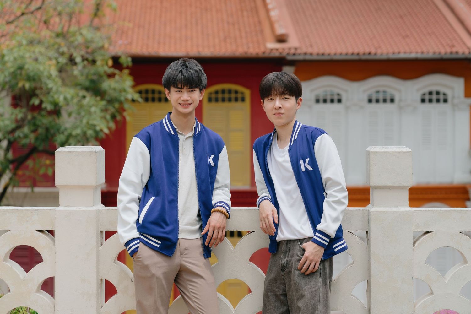 Two young men wearing matching blue and white varsity jackets pose together by a white fence near a red building.
