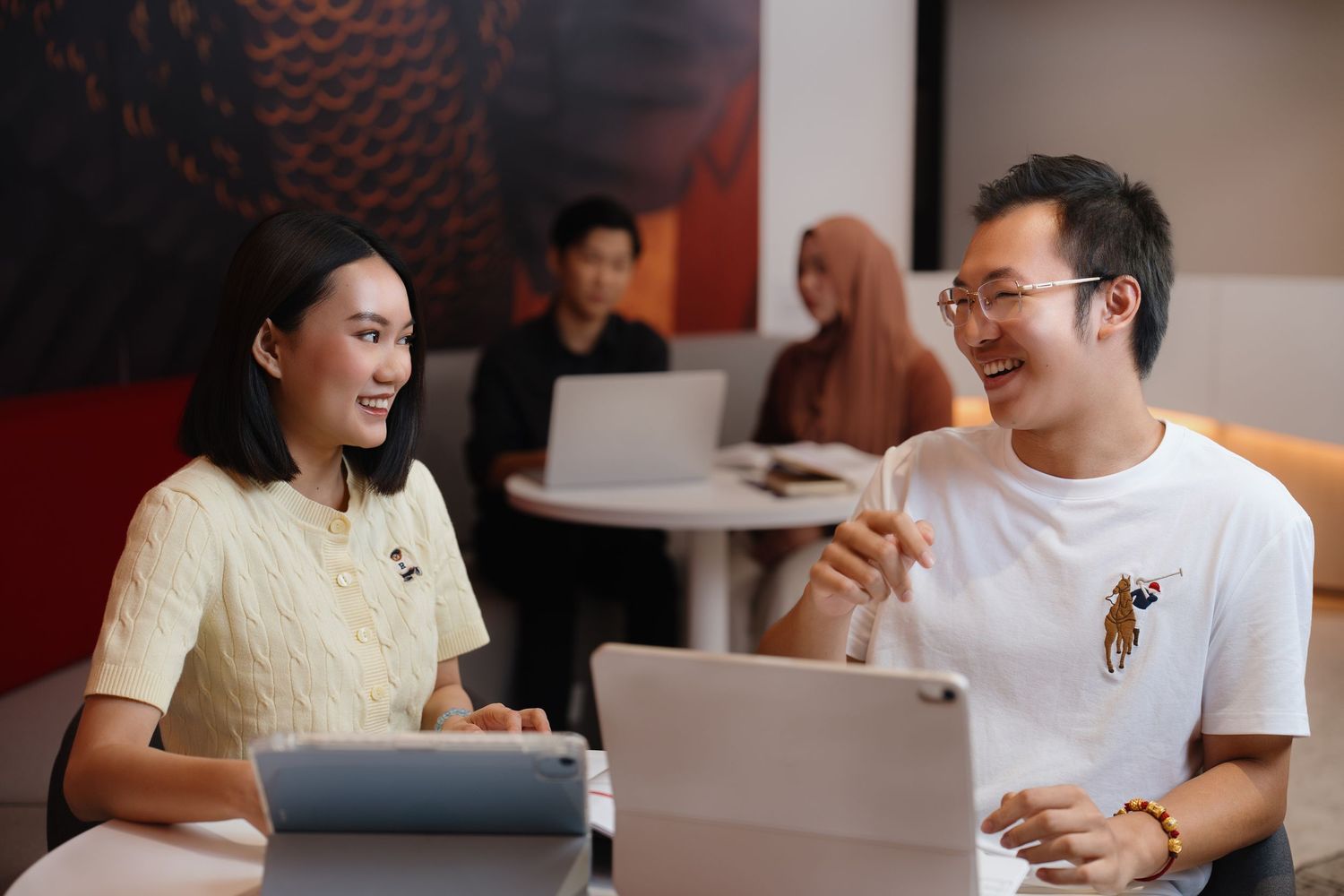 People in a cafe setting laugh and smile while working on laptops and having conversations at tables.