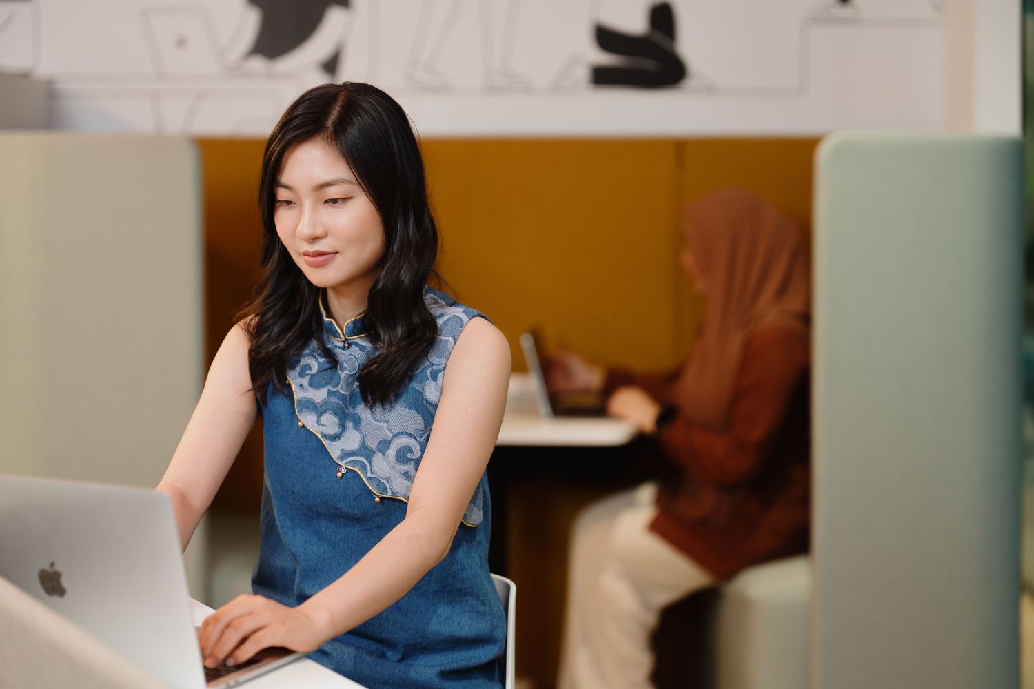 Professional in a denim top works at a desk with a laptop in a bright office setting.